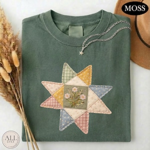 May include: A moss green t-shirt featuring a central star-shaped patchwork design. The star is composed of various patterned fabric segments in muted tones of yellow, blue, pink, and green, with a central square depicting a floral illustration.