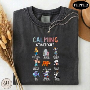 A black t-shirt with a colorful graphic design that reads "CALMING STRATEGIES" and features various calming activities such as giving oneself a hug, doing some stretches, drinking water, reading a book, and listening to music.