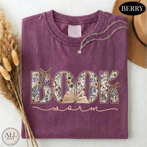 a purple t-shirt with the word "BOOK" embroidered on it, surrounded by floral designs. The t-shirt is placed on a white surface, with a hat and some dried flowers nearby.