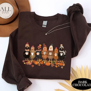 May include: A dark chocolate brown crewneck sweatshirt featuring a graphic print of seven chickens wearing colorful knit hats and scarves, each holding a white mug. The chickens stand among scattered autumn leaves and orange pumpkins.