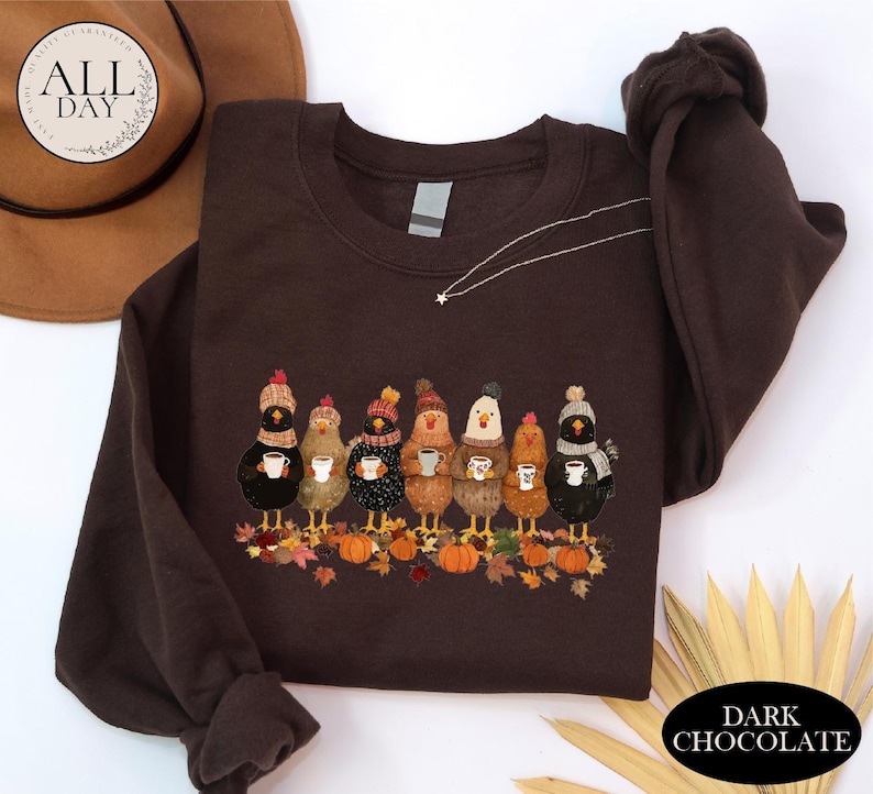 May include: A dark chocolate brown crewneck sweatshirt featuring a graphic print of seven chickens wearing colorful knit hats and scarves, each holding a white mug. The chickens stand among scattered autumn leaves and orange pumpkins.
