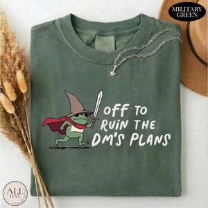 May include: Military green t-shirt featuring a cartoon frog wearing a wizard hat and cape, holding a sword. The text "off to ruin the DM's plans" is printed in white. A silver necklace with a star pendant is draped on the shirt.