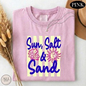 A pink t-shirt with the text "Sun, Salt & Sand" printed on it, along with illustrations of a sun, a seashell, and a sand dollar. The t-shirt is folded and placed on a white surface, with a brown hat and some dried flowers nearby.
