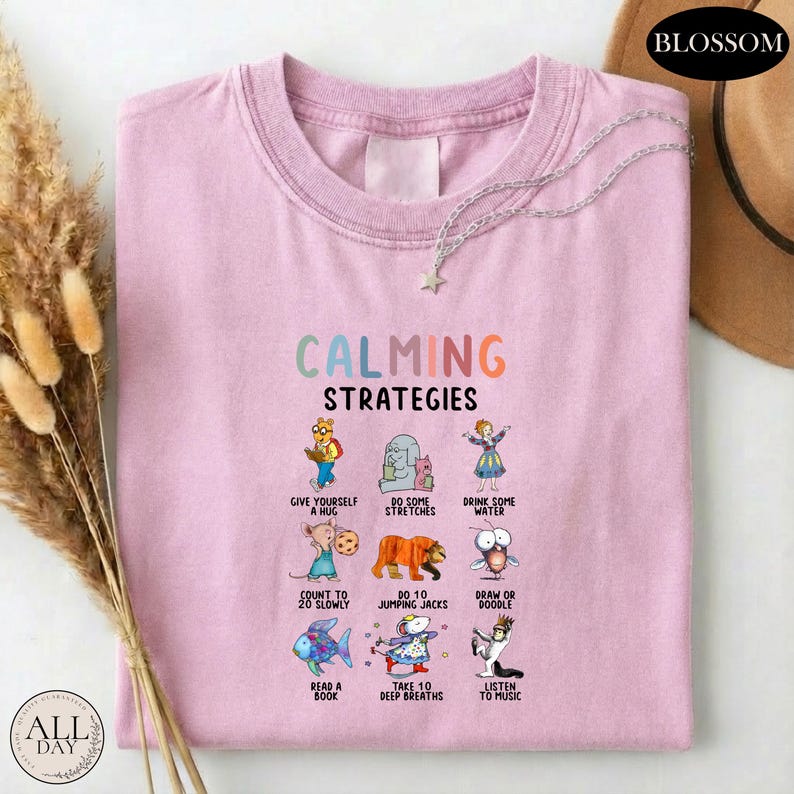 A pink t-shirt with a graphic design of various calming strategies and illustrations, including a bear, a bird, a cat, and other animals.