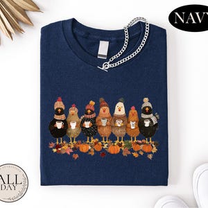 May include: A navy blue t-shirt featuring a graphic of seven chickens wearing winter hats and scarves, each holding a mug. The chickens stand among autumn leaves and pumpkins. A silver chain necklace is draped over the neckline of the shirt.