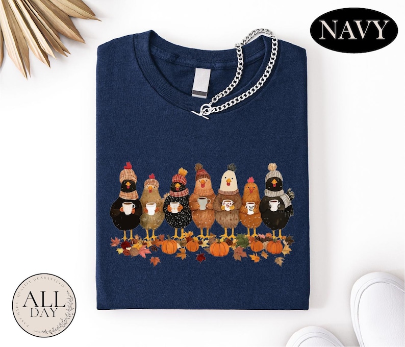 May include: A navy blue t-shirt featuring a graphic of seven chickens wearing winter hats and scarves, each holding a mug. The chickens stand among autumn leaves and pumpkins. A silver chain necklace is draped over the neckline of the shirt.