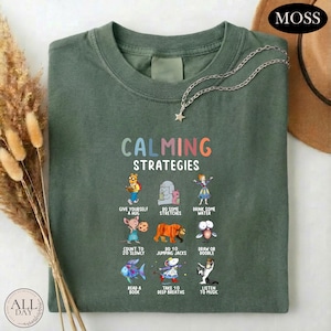 A green t-shirt with a colorful graphic design of various animals and objects, along with the text "Calming Strategies" printed on it. The t-shirt is laid out on a white surface, and there are some dried flowers and a hat visible in the background.