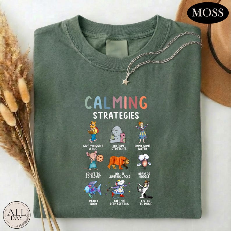 A green t-shirt with a colorful graphic design of various animals and objects, along with the text "Calming Strategies" printed on it. The t-shirt is laid out on a white surface, and there are some dried flowers and a hat visible in the background.