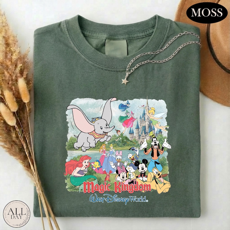 A green t-shirt with a colorful graphic of Disney characters from the Magic Kingdom, including Dumbo, Minnie Mouse, Donald Duck, and other iconic figures, along with a castle and a castle-like structure.