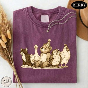 a purple t-shirt with a graphic design of a bear, cat, duck, chicken, and owl, all wearing party hats.