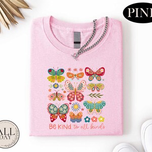 A pink t-shirt with a colorful butterfly design and the text "Be kind to all kinds" printed on it, along with a chain and a pair of white sneakers.