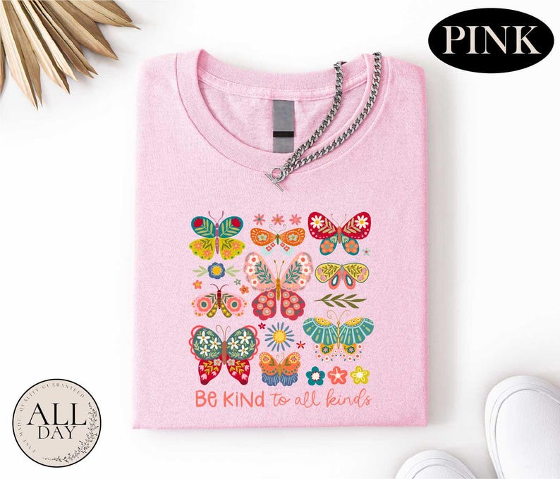 A pink t-shirt with a colorful butterfly design and the text "Be kind to all kinds" printed on it, along with a chain and a pair of white sneakers.