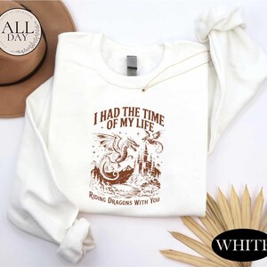 a white long-sleeved shirt with a graphic design of a dragon and castle, along with the text "I HAD THE TIME OF MY LIFE RIDING DRagons WITH YOU". The shirt is laid out on a white background, and there is a brown hat and some straws in the background.