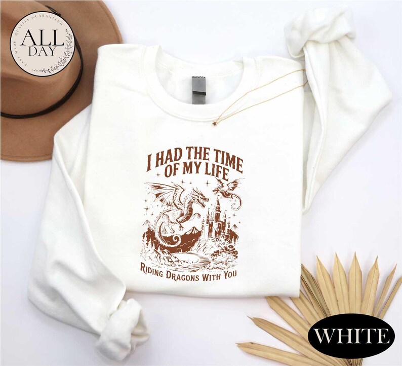 a white long-sleeved shirt with a graphic design of a dragon and castle, along with the text "I HAD THE TIME OF MY LIFE RIDING DRagons WITH YOU". The shirt is laid out on a white background, and there is a brown hat and some straws in the background.