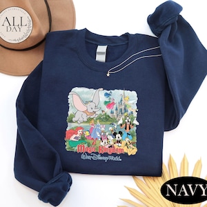 a navy blue sweatshirt with a Disney-themed graphic, including characters from the Disney film "Magic Kingdom". The sweatshirt is placed on a white surface, with a brown hat and a yellow flower nearby.