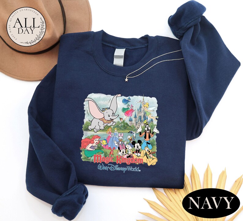 a navy blue sweatshirt with a Disney-themed graphic, including characters from the Disney film "Magic Kingdom". The sweatshirt is placed on a white surface, with a brown hat and a yellow flower nearby.
