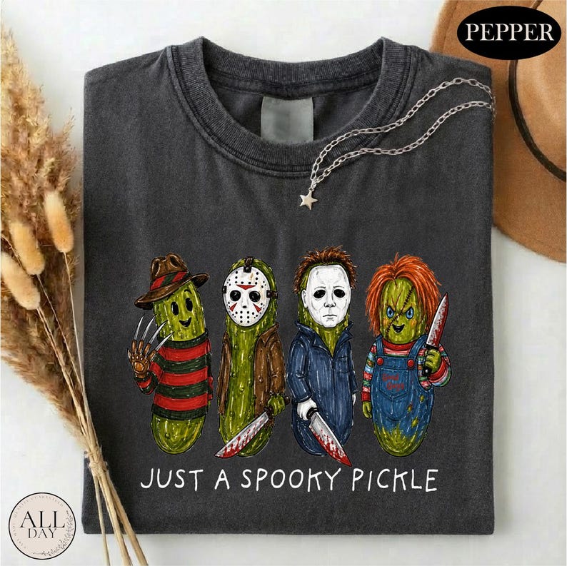May include: A dark gray t-shirt featuring a graphic of four green pickles dressed as horror movie characters. Below the characters, the text reads JUST A SPOOKY PICKLE. The shirt includes a silver-toned chain necklace with a small star charm.