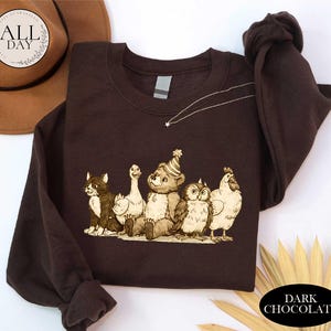 a brown t-shirt with a cartoon illustration of a bear, cat, duck, chicken, and owl, all wearing party hats. The t-shirt is laid out on a white surface, with a brown hat and some leaves visible in the background.