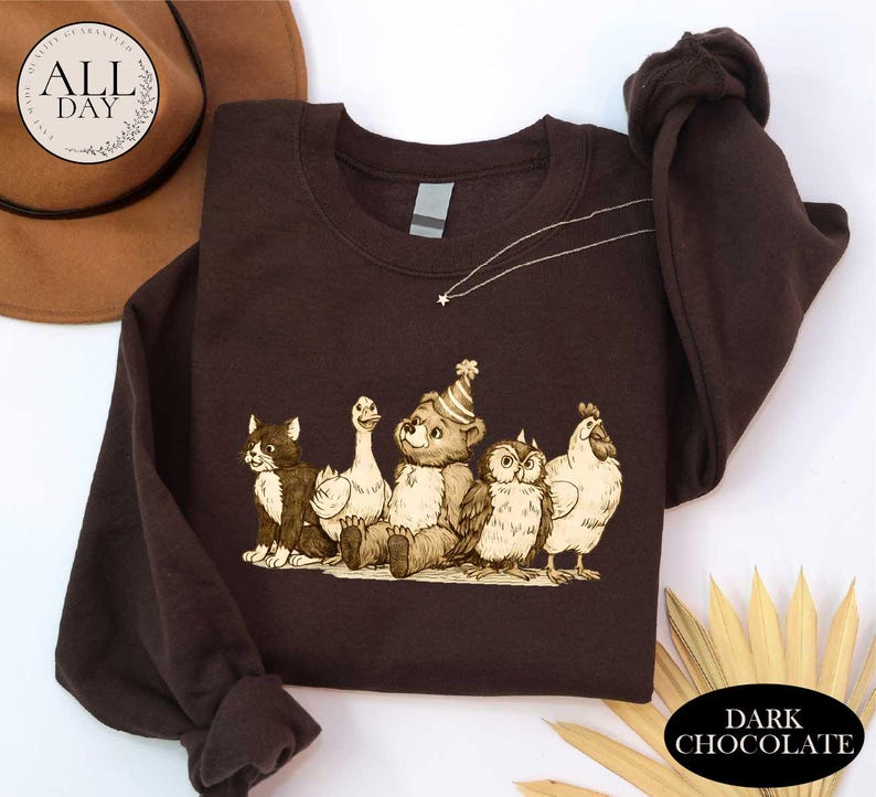 a brown t-shirt with a cartoon illustration of a bear, cat, duck, chicken, and owl, all wearing party hats. The t-shirt is laid out on a white surface, with a brown hat and some leaves visible in the background.