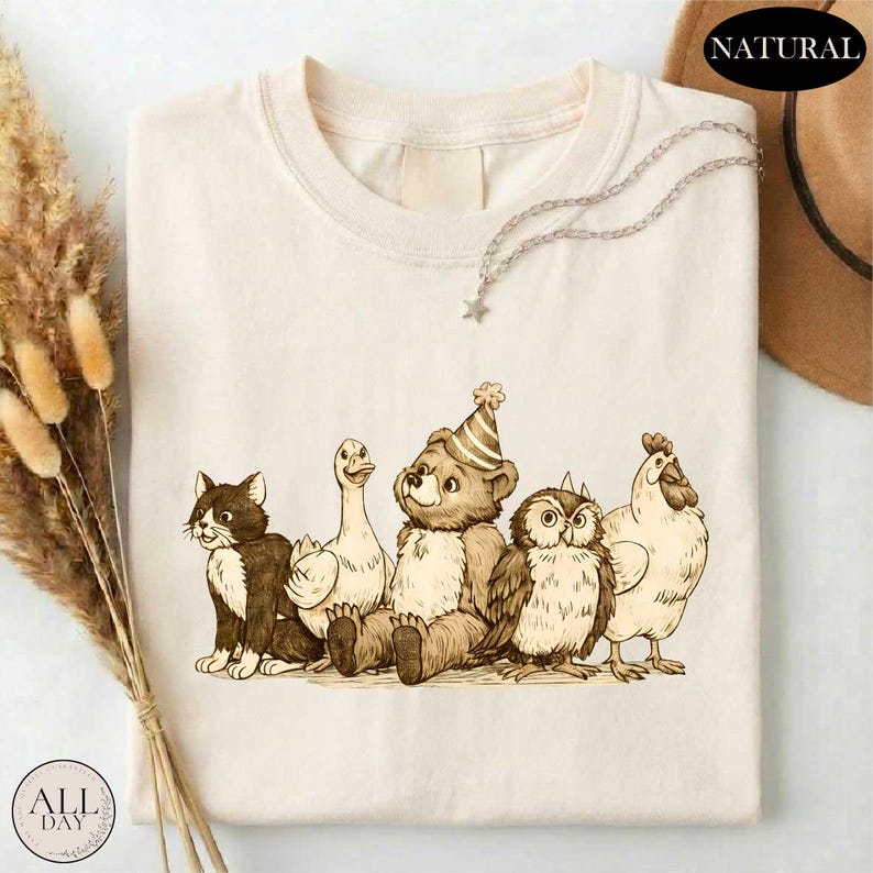 A t-shirt with a cartoon illustration of a bear, cat, duck, chicken, and owl, all wearing party hats, sitting together.
