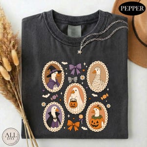 May include: A charcoal gray t-shirt featuring a graphic print of five geese in oval frames, dressed in Halloween costumes including a witch, a mummy, a ghost, a vampire, and a pumpkin. The design includes purple and orange bows and candy.