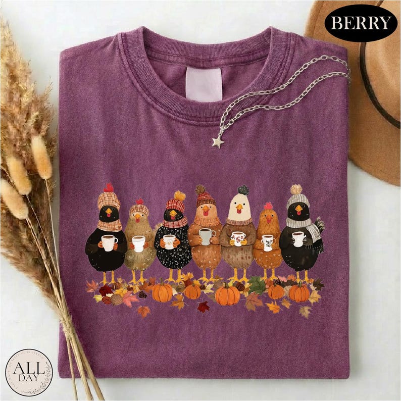 May include: A berry-colored t-shirt featuring a graphic of seven chickens wearing knit hats and scarves, each holding a white mug. The chickens stand on a pile of autumn leaves and pumpkins. A silver star-shaped pendant necklace rests on the shirt.