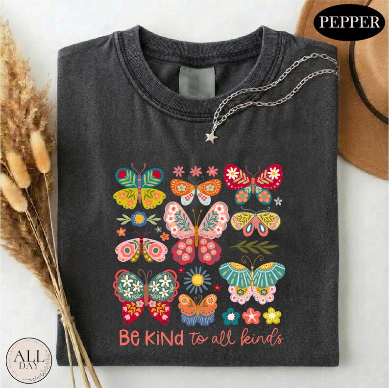 a black t-shirt with a colorful graphic design of butterflies and flowers, along with the text "Be Kind to all kinds" printed on it. The t-shirt is laid out on a white surface, and there is a brown hat and some dried flowers in the background.