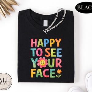 a black t-shirt with a colorful text design that says "HAPPY TO SEE YOUR FACE" and includes a flower and a smiley face.