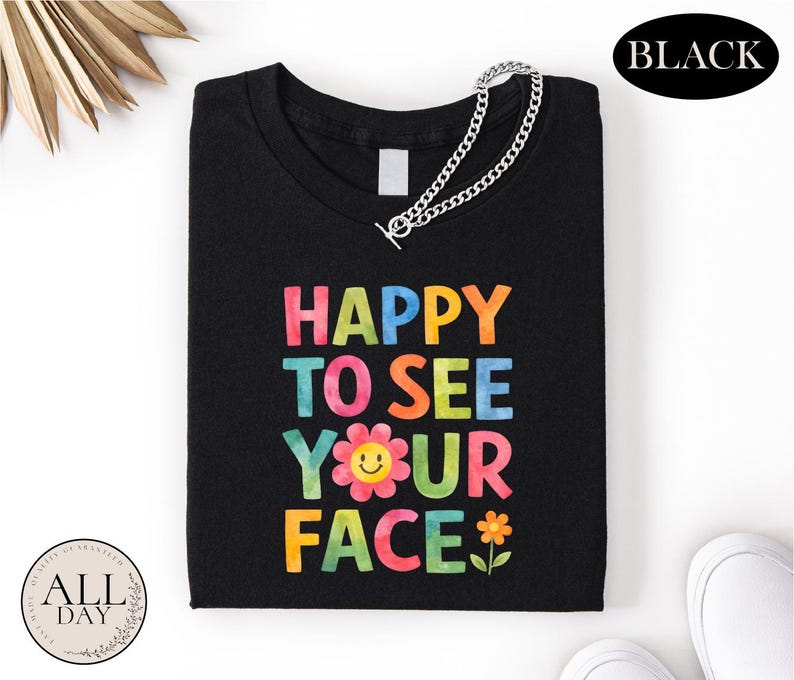 a black t-shirt with a colorful text design that says "HAPPY TO SEE YOUR FACE" and includes a flower and a smiley face.