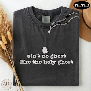 May include: A charcoal gray crew neck t-shirt featuring a small white ghost graphic and the text "ain't no ghost like the holy ghost" printed in a distressed white font. A delicate silver-toned chain necklace with a small star charm rests on the collar.