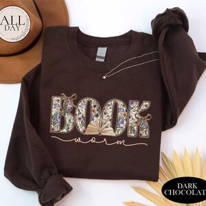 a brown sweatshirt with the word "BOOK" embroidered on it, along with the word "Worm" written below it. The sweatshirt is placed on a white surface, and there is a brown hat and some gold leaves in the background.