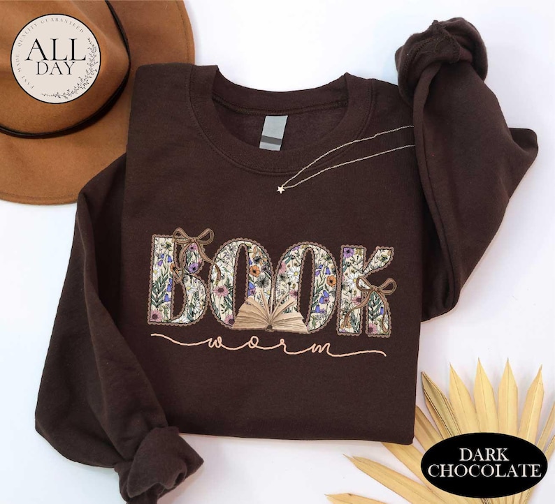 a brown sweatshirt with the word "BOOK" embroidered on it, along with the word "Worm" written below it. The sweatshirt is placed on a white surface, and there is a brown hat and some gold leaves in the background.