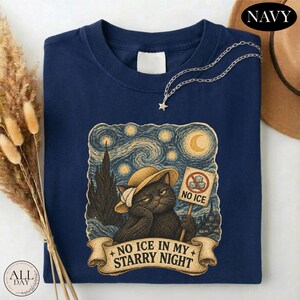 May include: A navy blue t-shirt featuring a graphic of a black cat wearing a straw hat, holding a sign that says NO ICE with a crossed-out ice cube icon. Below the cat, a banner displays the text NO ICE IN MY STARRY NIGHT in a swirling sky.
