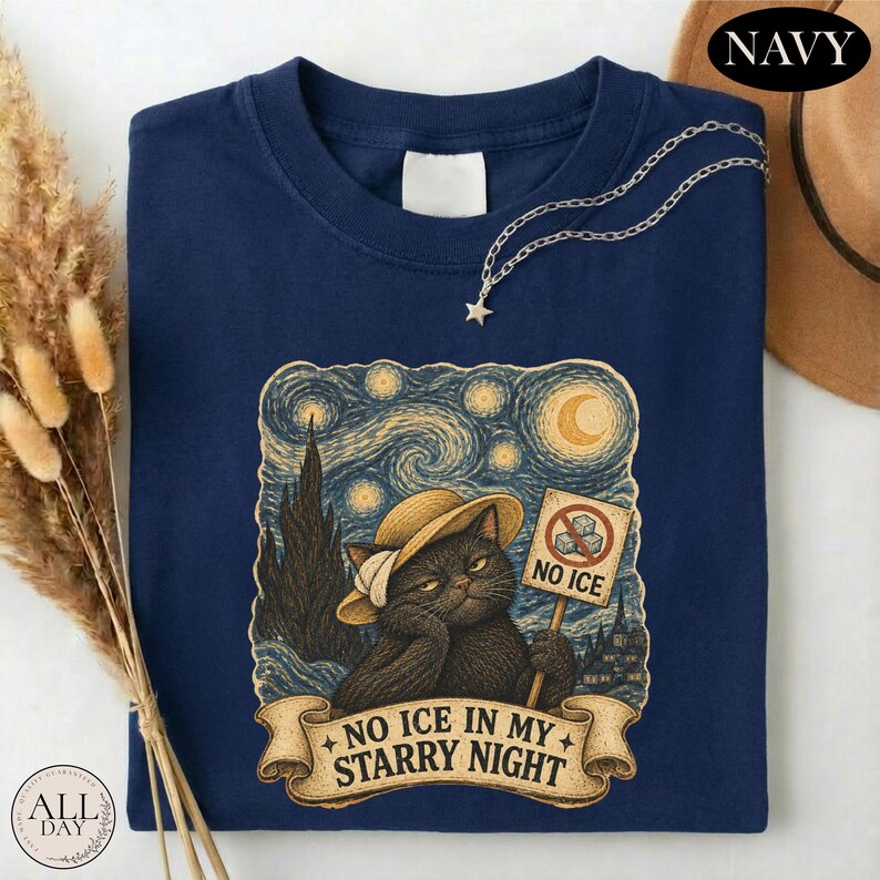 May include: A navy blue t-shirt featuring a graphic of a black cat wearing a straw hat, holding a sign that says NO ICE with a crossed-out ice cube icon. Below the cat, a banner displays the text NO ICE IN MY STARRY NIGHT in a swirling sky.