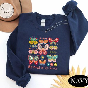 A navy blue sweatshirt with a colorful butterfly design and the text "Be Kind to all kinds" printed on it, along with a brown hat and some straw.