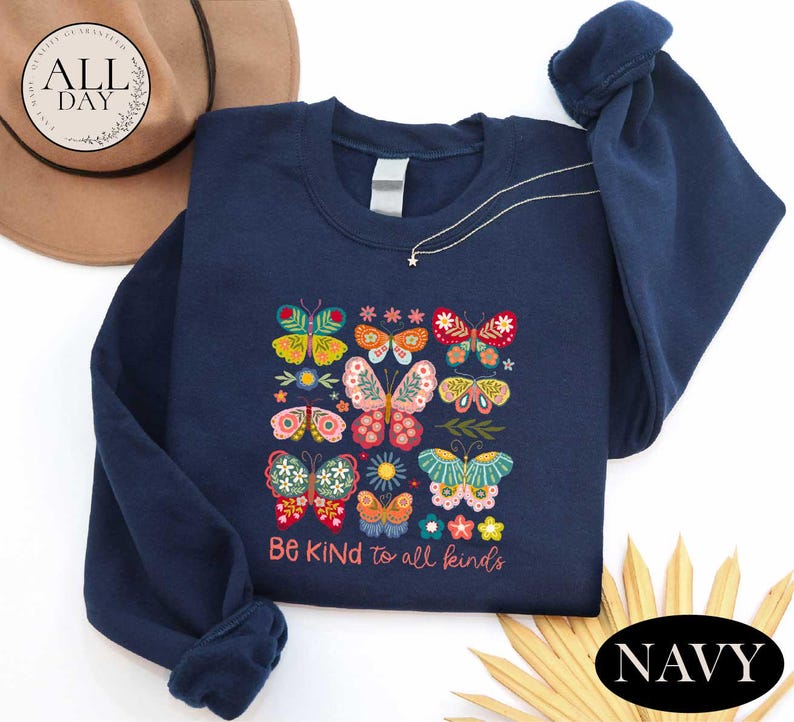 A navy blue sweatshirt with a colorful butterfly design and the text "Be Kind to all kinds" printed on it, along with a brown hat and some straw.