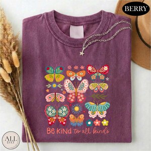 a purple t-shirt with a colorful butterfly design and the text "Be Kind to all kinds" printed on it. The t-shirt is folded and placed on a white surface, with a brown hat and dried flowers nearby.