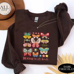 a brown t-shirt with a colorful butterfly design and the text "Be Kind to all Kinds" printed on it. The t-shirt is laid out on a white surface, with a brown hat and some leaves visible in the background.