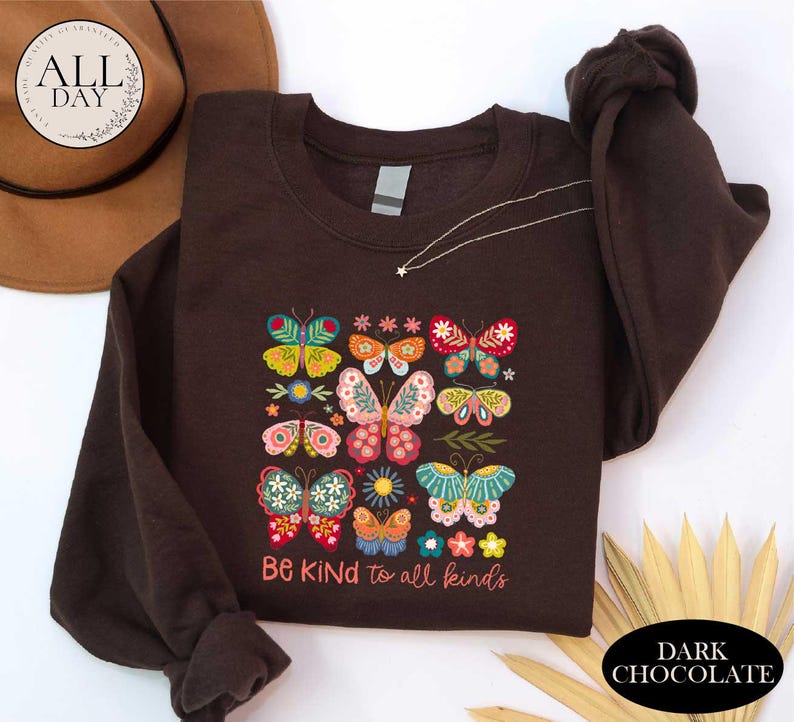 a brown t-shirt with a colorful butterfly design and the text "Be Kind to all Kinds" printed on it. The t-shirt is laid out on a white surface, with a brown hat and some leaves visible in the background.