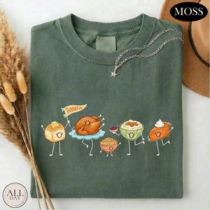 May include: A dark olive green t-shirt featuring a graphic of five anthropomorphic Thanksgiving food items including a dinner roll, a roasted turkey holding a flag that says THANKFUL, stuffing, mashed potatoes, and a slice of pumpkin pie.