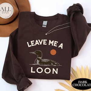 A brown sweatshirt with a graphic of a duck and the text "LEAVE ME A LON" printed on it, along with a hat and some leaves.