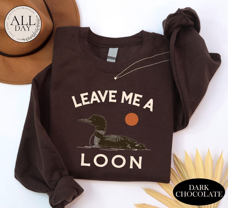 A brown sweatshirt with a graphic of a duck and the text "LEAVE ME A LON" printed on it, along with a hat and some leaves.