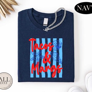 a navy blue t-shirt with the text "Tacos & Mangos" printed on it, along with a graphic of a blue and white striped pattern and a seashell design. The t-shirt is displayed on a white background, and there are some other items, such as a chain and a pair of white shoes, visible in the image.