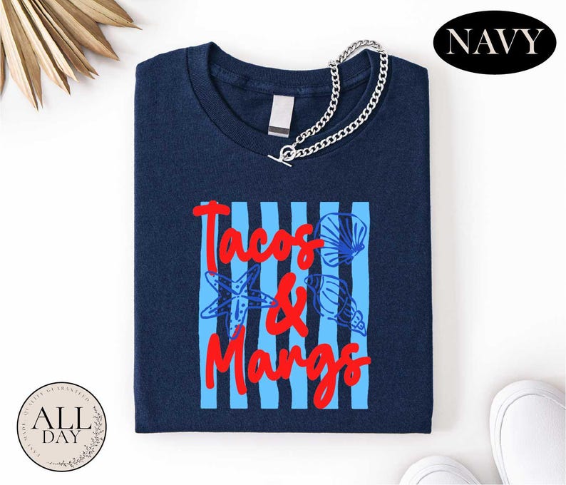 a navy blue t-shirt with the text "Tacos & Mangos" printed on it, along with a graphic of a blue and white striped pattern and a seashell design. The t-shirt is displayed on a white background, and there are some other items, such as a chain and a pair of white shoes, visible in the image.