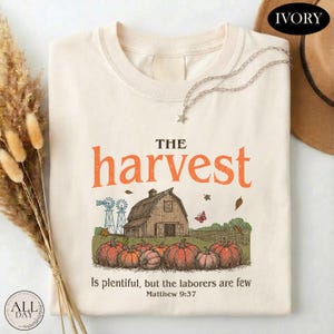 May include: An ivory t-shirt featuring a graphic of a barn, pumpkins, and windmills. The text reads: THE harvest Is plentiful, but the laborers are few Matthew 9:37. The design includes orange, brown, and green colors on a light-colored fabric.