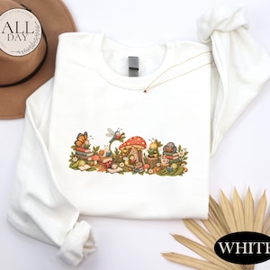 K&ouml;nnte beinhalten: Ein wei&szlig;es Langarm-Sweatshirt mit einem farbenfrohen Grafikdruck verschiedener Insekten, darunter ein Schmetterling, ein Marienk&auml;fer und eine Biene, die B&uuml;cher um einen gro&szlig;en roten Pilz herum lesen, erg&auml;nzt durch kleine Blumen und gr&uuml;ne Bl&auml;tter.