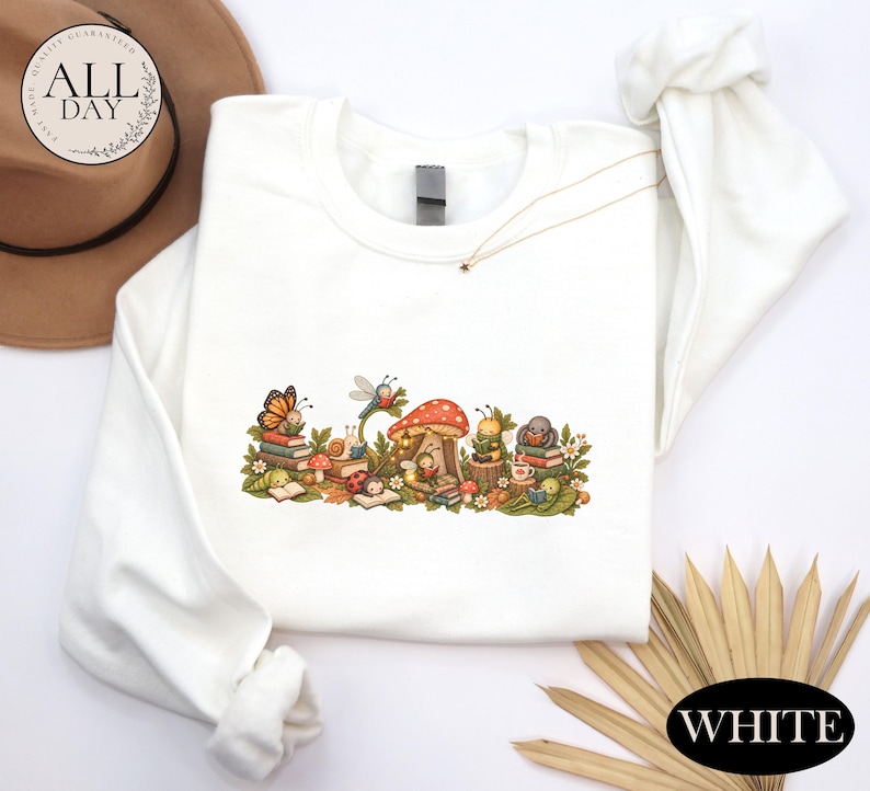 K&ouml;nnte beinhalten: Ein wei&szlig;es Langarm-Sweatshirt mit einem farbenfrohen Grafikdruck verschiedener Insekten, darunter ein Schmetterling, ein Marienk&auml;fer und eine Biene, die B&uuml;cher um einen gro&szlig;en roten Pilz herum lesen, erg&auml;nzt durch kleine Blumen und gr&uuml;ne Bl&auml;tter.