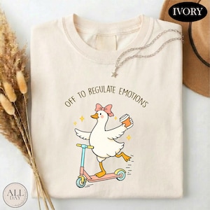 May include: Ivory-colored t-shirt featuring a cartoon goose riding a pink scooter and holding a coffee cup. The goose wears a pink bow and the text "OFF TO REGULATE EMOTIONS" is printed above the goose. A silver necklace with a star pendant is draped on the shirt.