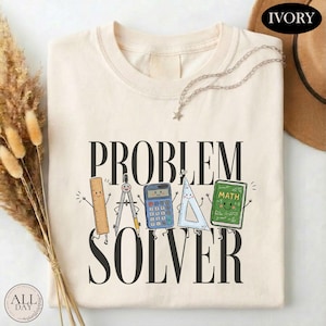 May include: An ivory t-shirt featuring the black text "PROBLEM SOLVER" and a graphic of a smiling ruler, compass, calculator, triangle, and a green book labeled "MATH". The design includes small arms and legs on each school supply item.