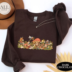 K&ouml;nnte beinhalten: Ein dunkelbraunes Sweatshirt mit Rundhalsausschnitt und einem farbenfrohen Grafikdruck, der verschiedene Insekten und kleine Lebewesen zeigt, die zwischen Pilzen, Bl&auml;ttern und Blumen B&uuml;cher lesen. Das Design umfasst einen Schmetterling, eine Schnecke, einen Marienk&auml;fer und eine Biene.